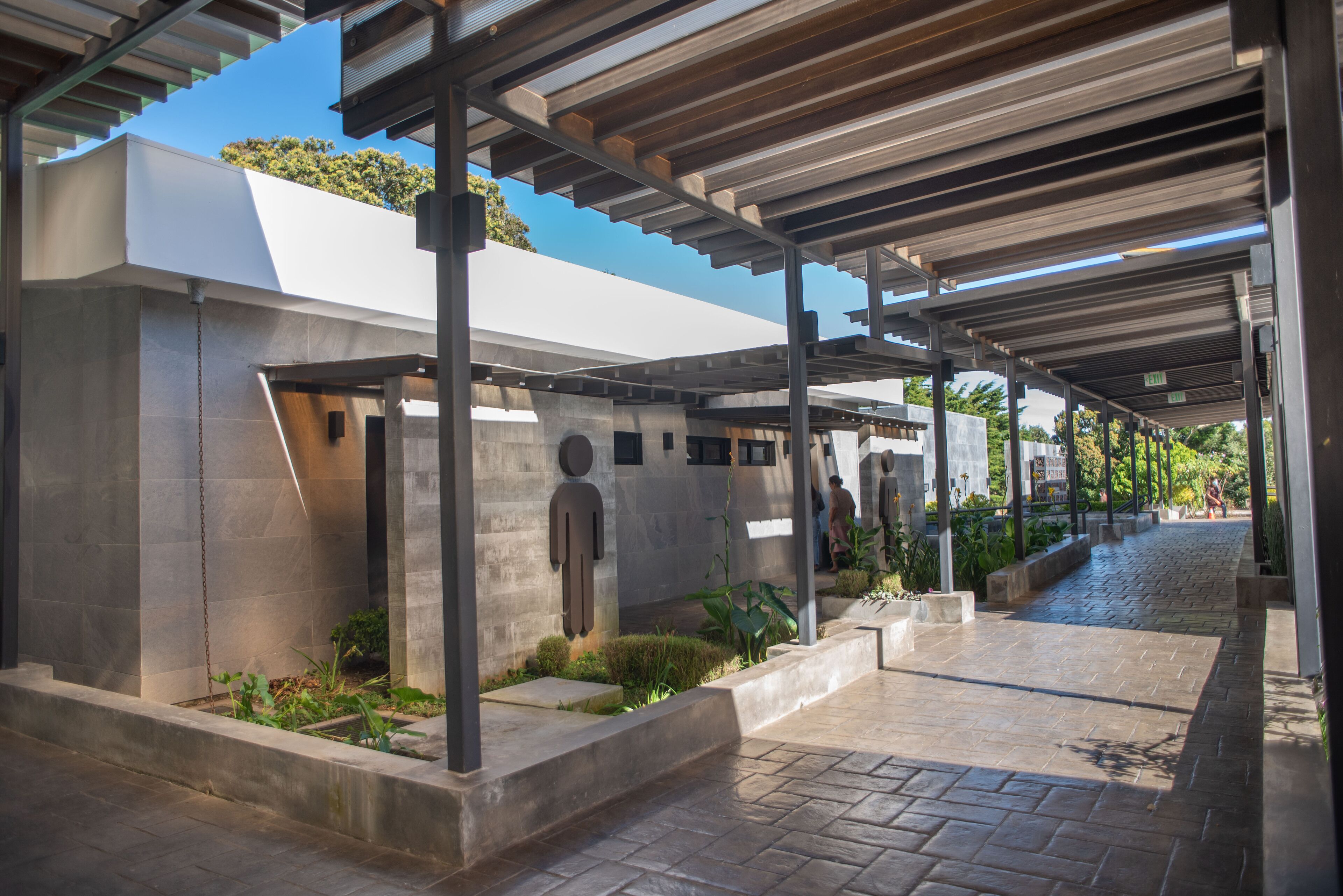 A covered outdoor area with a modern architectural design, featuring a concrete structure, metal beams, and a paved walkway.