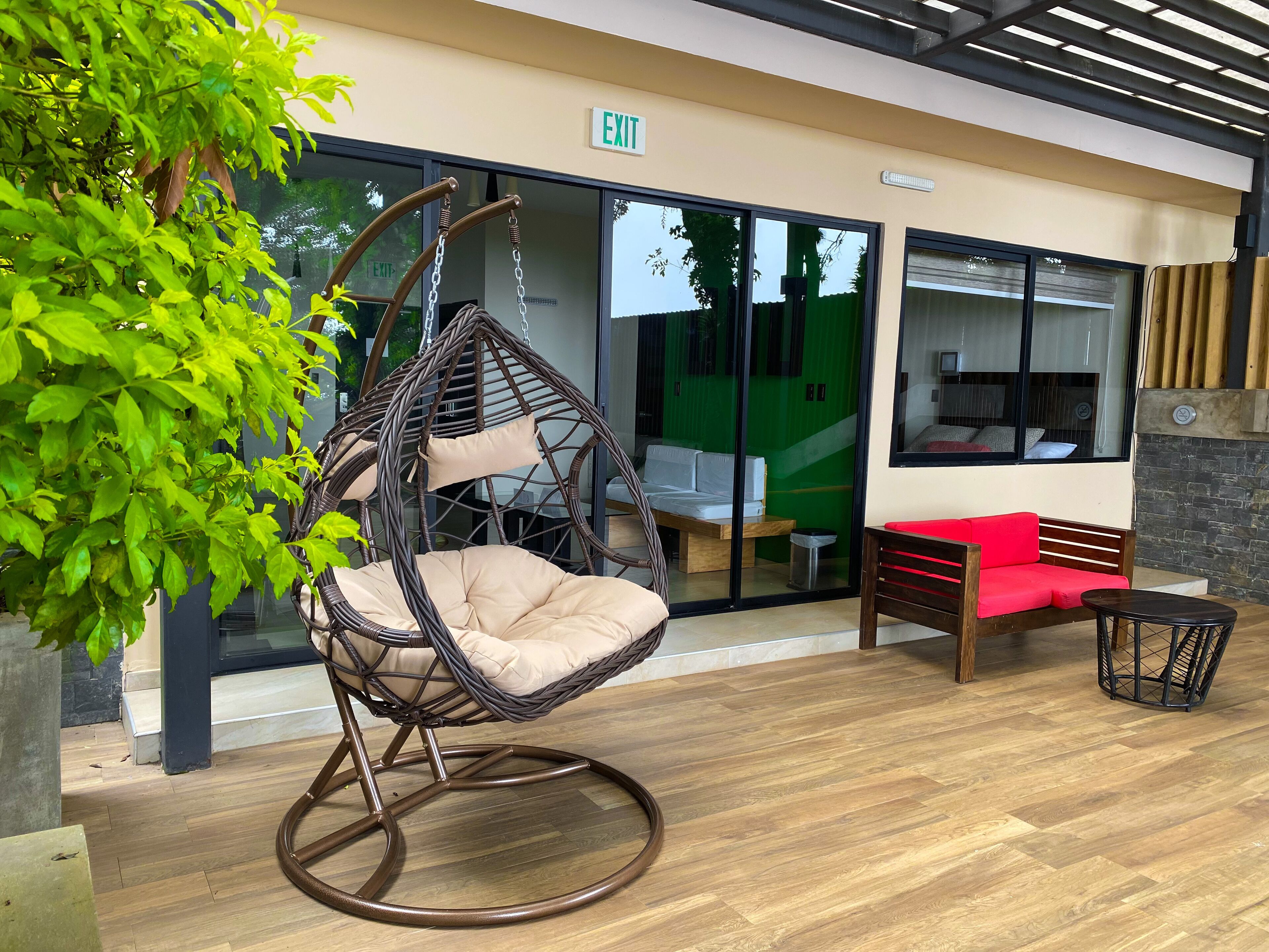 A wicker hanging chair with a cushion, a red sofa, and a wooden bench in an indoor area with a wooden floor and glass doors leading to a green room.