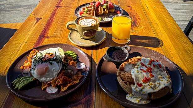 Una mesa de madera con platos de desayuno, una taza de café y un vaso de zumo de naranja, con vistas a un paisaje escénico que incluye una montaña y abundante vegetación.