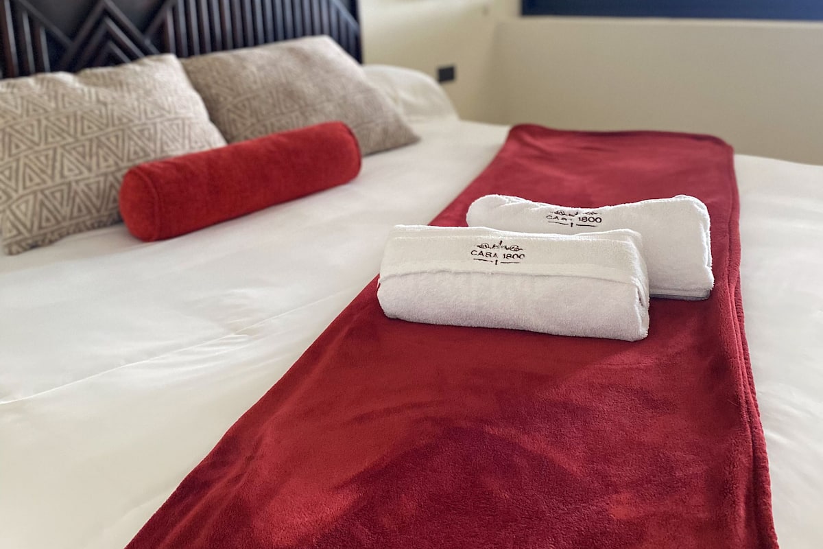 Una cama bien hecha con una manta roja, almohadas blancas y un cojín rojo. Hay dos toallas blancas dobladas sobre la cama.