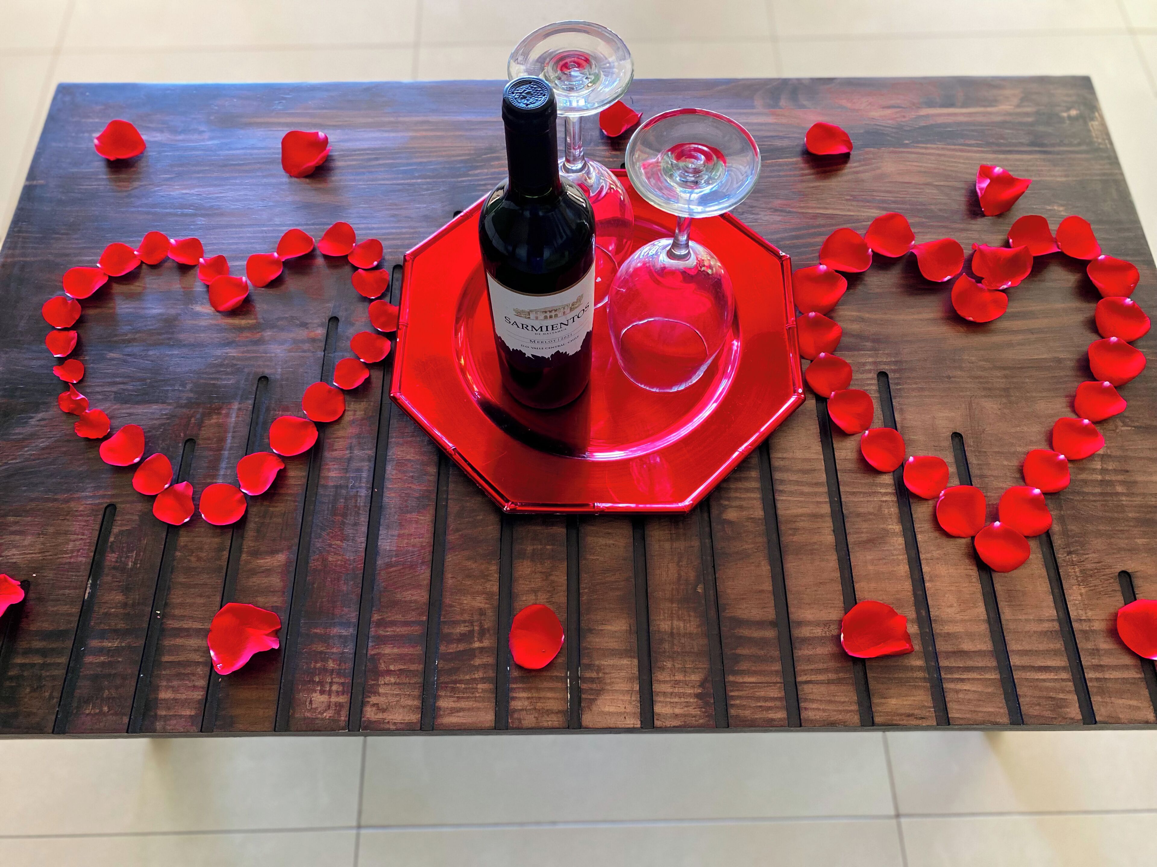 A wooden table with a heart-shaped arrangement of red rose petals, a bottle of wine, and two wine glasses.