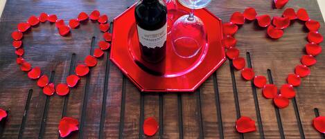 A wooden table with a heart-shaped arrangement of red rose petals, a bottle of wine, and two wine glasses.