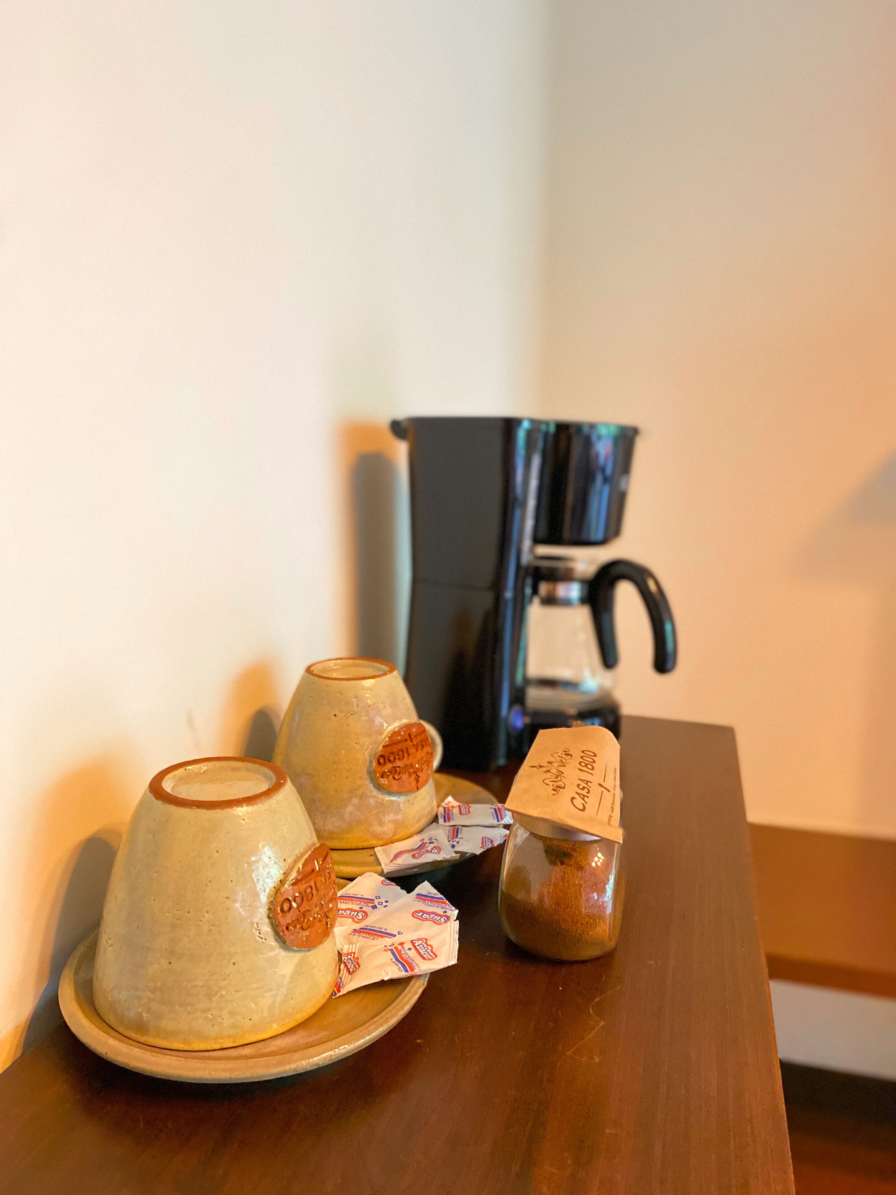 A table with ceramic cups, a plate, and a jar of sugar, with a coffee machine in the background.