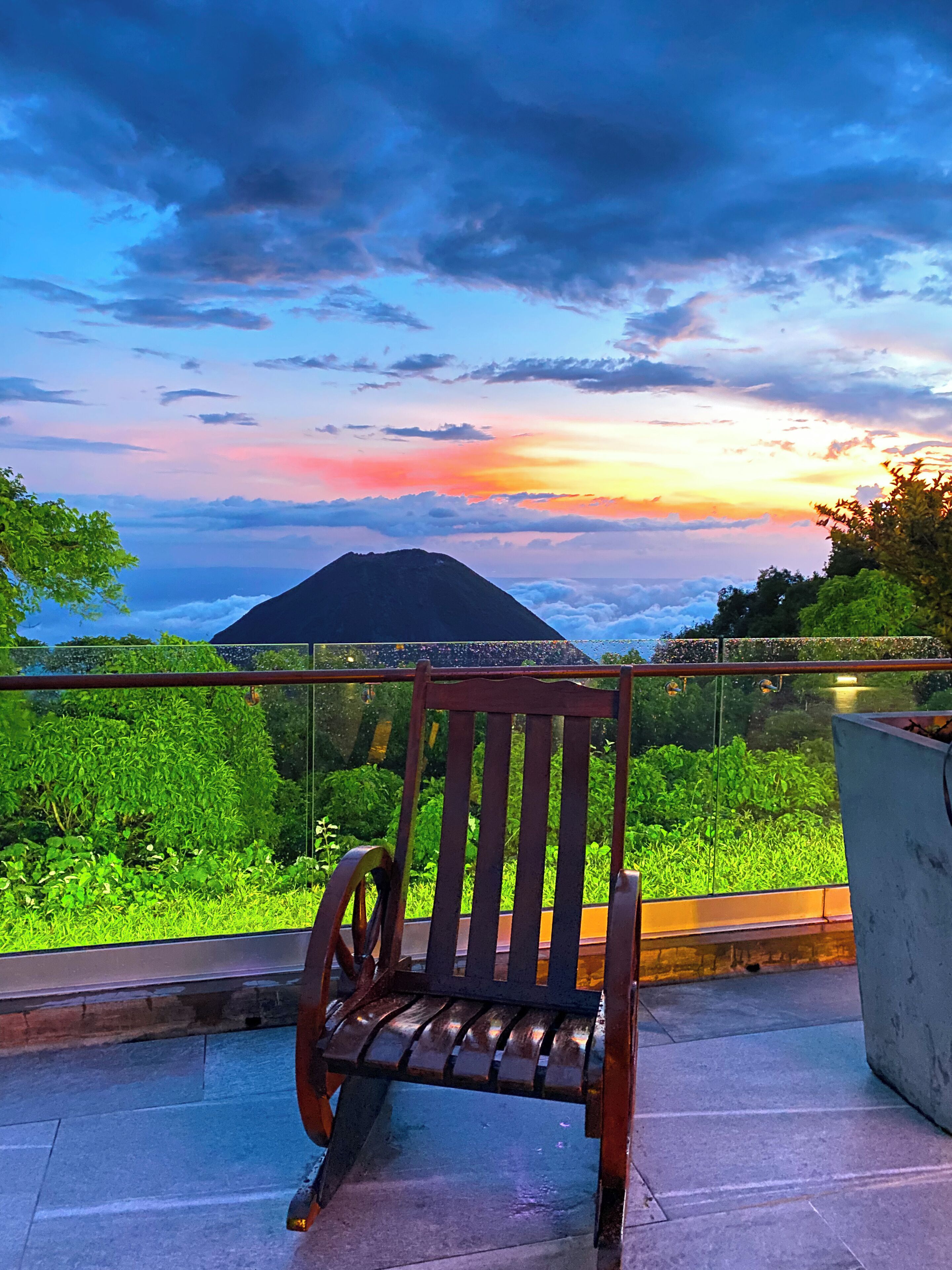 A wooden bench on a balcony overlooking a lush green landscape and a mountain during sunset.