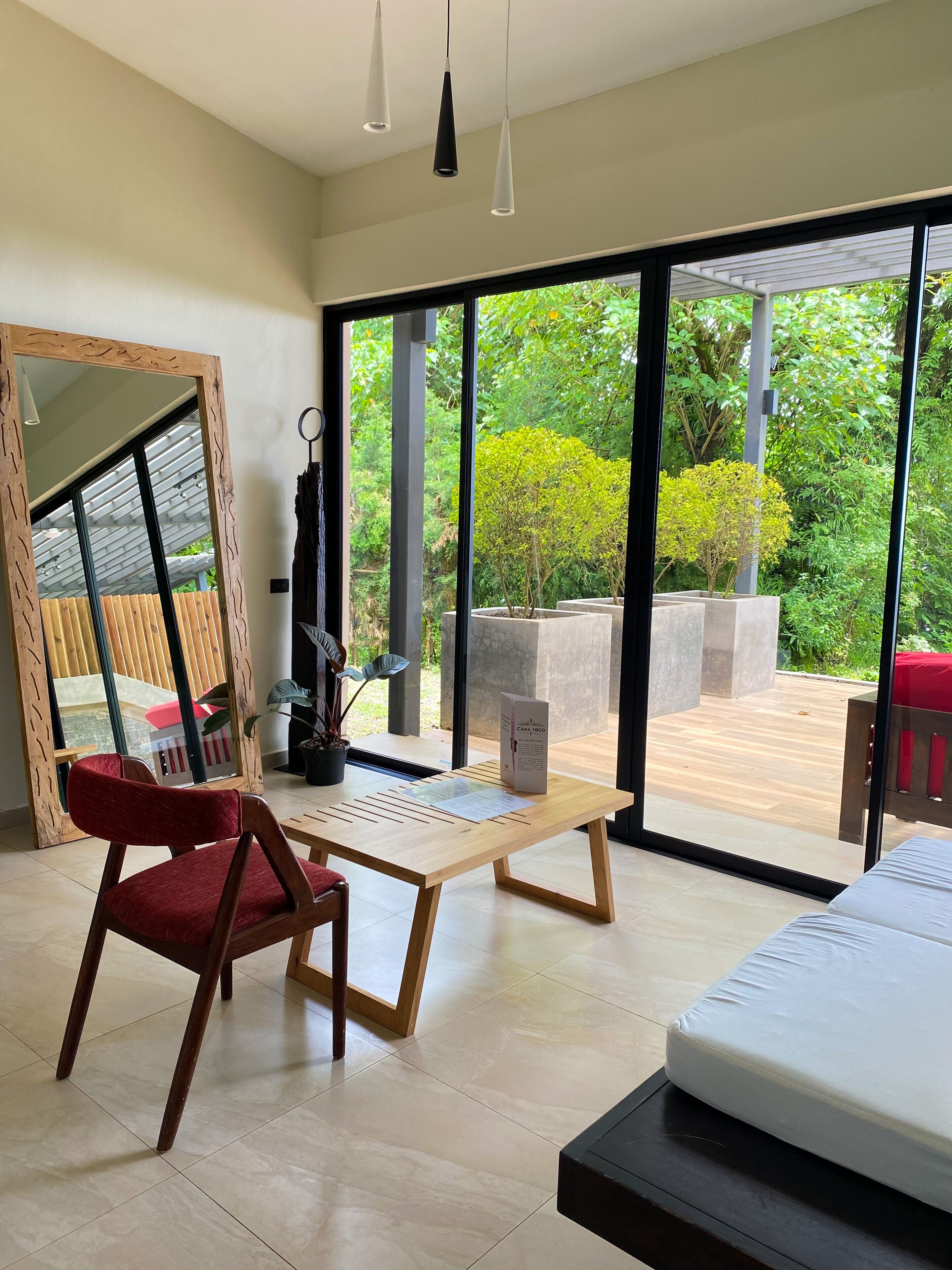 Il s’agit d’une chambre moderne dotée d’une grande porte-fenêtre coulissante qui donne sur un patio extérieur. À l’intérieur, on trouve une table en bois avec une chaise rouge, un lit et un miroir avec un cadre en bois.