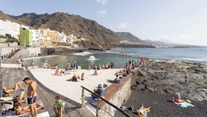 A beach with people sunbathing and swimming, a rocky shoreline, and buildings in the background. - Home2Book SeaFront Studio Punta del Hidalgo (Punta del Hidalgo)