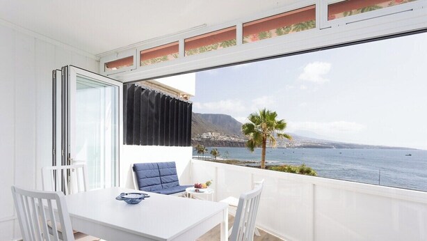 A balcony with a white table and chairs, a view of the sea and mountains, and a palm tree. - Home2Book SeaFront Studio Punta del Hidalgo (Punta del Hidalgo)