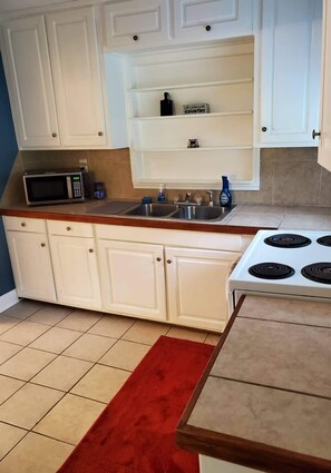 Compact kitchen with a microwave, sink, and stove, featuring two shelves above the sink.