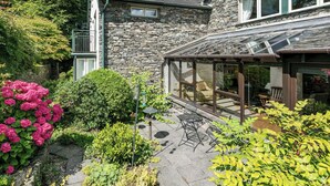 Une maison en pierre dotée d’une véranda en verre, entourée d’une végétation luxuriante et de plantes en fleurs.