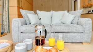 Un plateau de petit-déjeuner avec des croissants, du café et du jus d’orange, posé sur une table en bois dans un salon doté d’un canapé gris et de coussins à motifs.