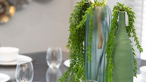 Et spisebord dækket med hvide tallerkener, skåle og klare vinglas. Der er to grønne vaser med planter på bordet.
