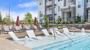 A pool area with lounge chairs and umbrellas, a stone wall, and a multi-story building in the background. - Luxury Apt | Work from here | King Bed | Fast WiFi |6mi to CLT✈️ (Charlotte)