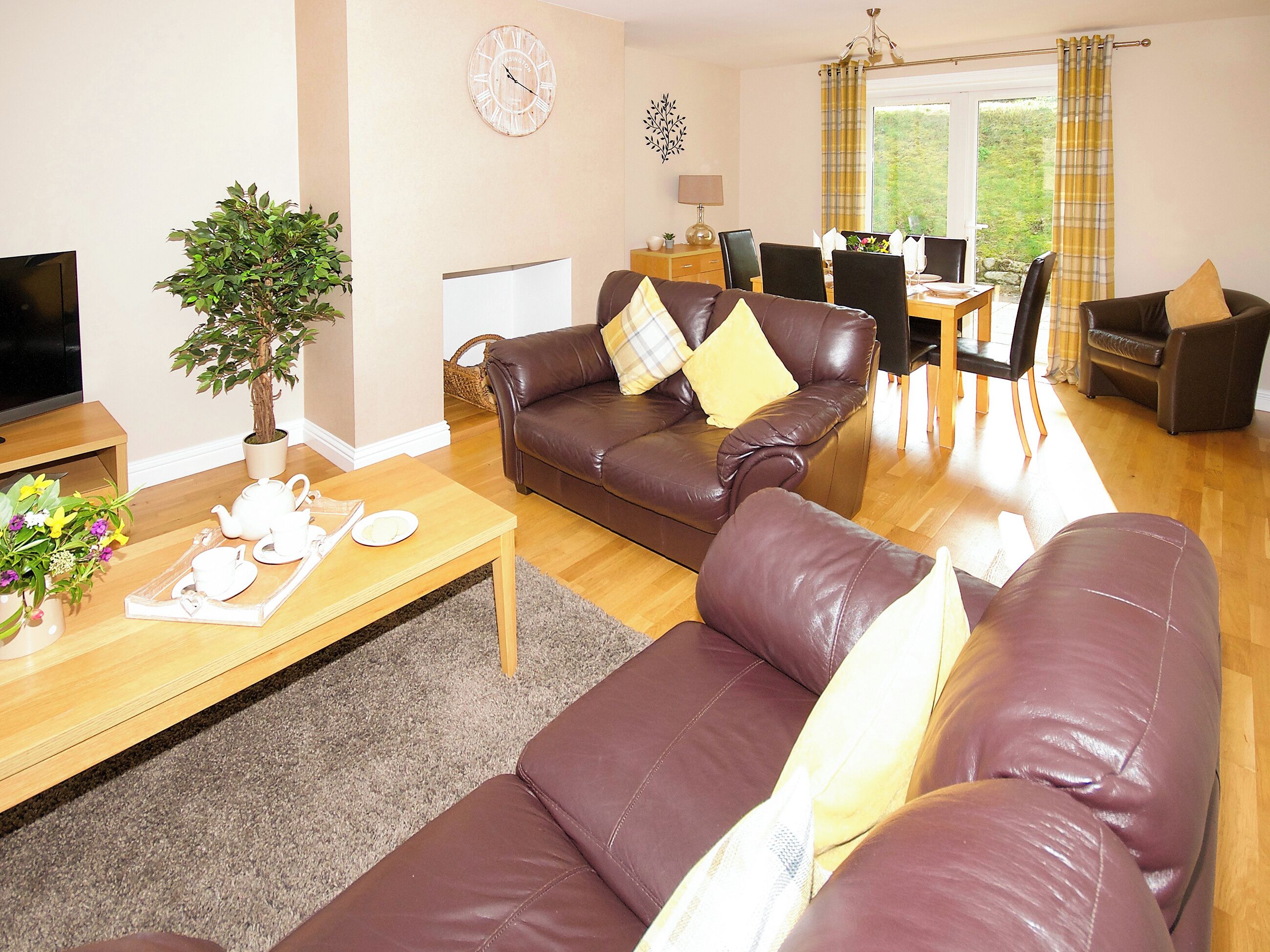 A living room with a brown leather sofa, a wooden coffee table, a TV, a potted plant, and a dining area with a table and chairs.