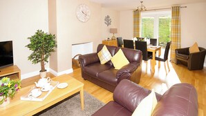 A living room with a brown leather sofa, a wooden coffee table, a TV, a potted plant, and a dining area with a table and chairs. - Single storey cottage for 4/6 in Twynholm only 3 miles from Kirkcudbright. (Twynholm by Kirkcudbright)