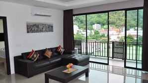A modern living room with a black leather sofa, a coffee table, and a large window offering a view of a cityscape and greenery. - Buasri Boutique Patong (Patong)
