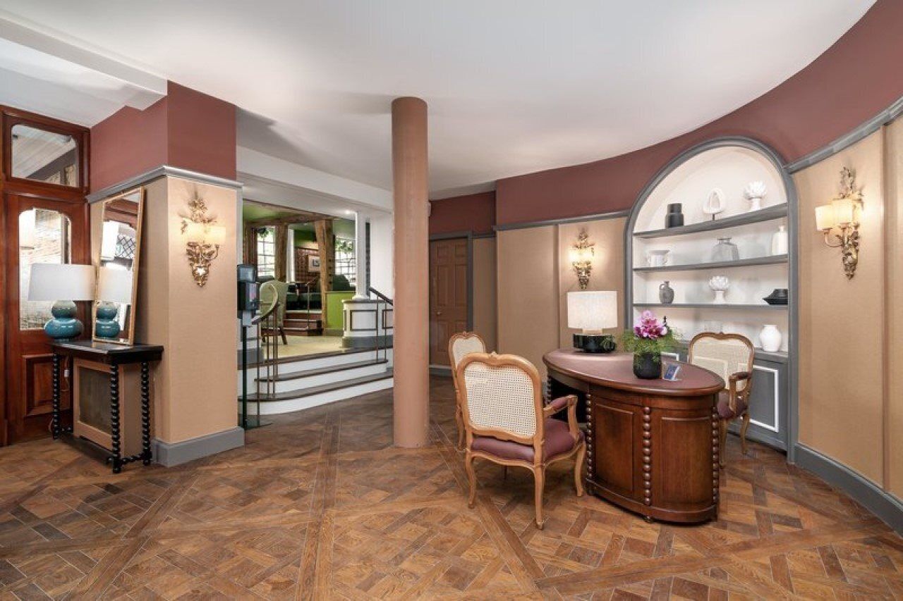 A hotel lobby with a wooden reception desk, a seating area with a chair, and a shelving unit with decorative items.