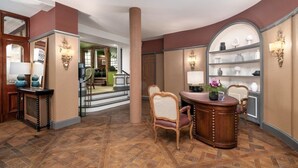 Un hall de hotel con un mostrador de recepción de madera, una zona de estar con una silla y un estante con objetos decorativos.