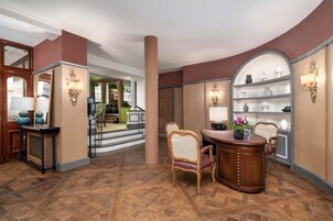 A hotel lobby with a wooden reception desk, a seating area with a chair, and a shelving unit with decorative items.