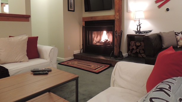A cozy living room with a fireplace, a television mounted above it, and a sofa with red cushions. - Best Location! Renovated center village Okemo 2 BR - free shuttle outside door! (Ludlow)