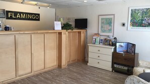 A reception desk with a sign reading "FLAMINGO", a television mounted on the wall, a framed picture, and a small plant on the counter.