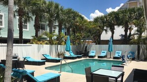 A pool area with lounge chairs and a table, surrounded by palm trees and a white fence.