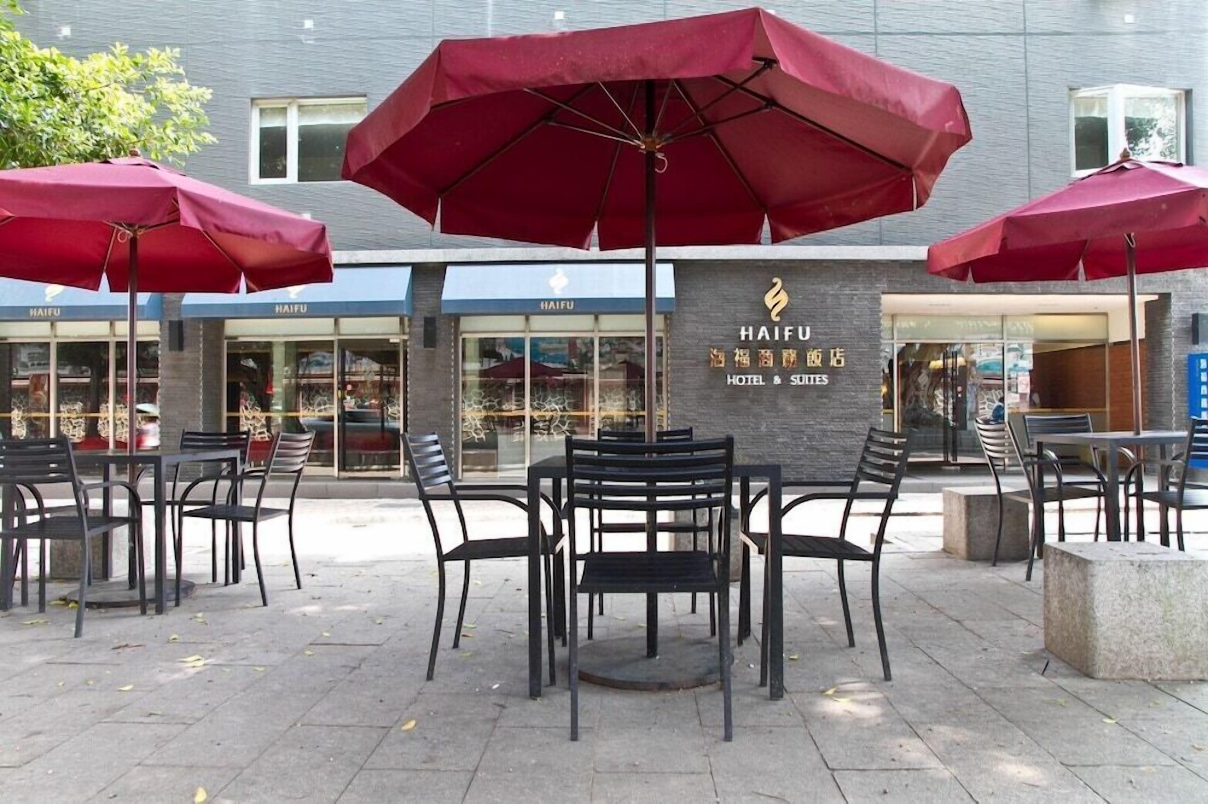 An outdoor seating area with tables and chairs, red umbrellas, and a building with 'HAIFU HOTEL & SITES' signage.