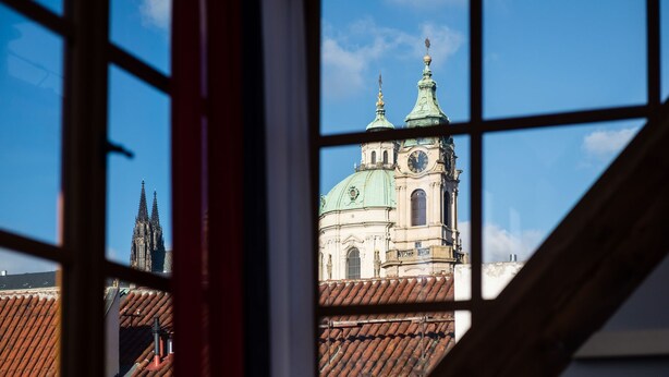 A view of a church with a clock tower and a spire through a window with a grid pattern. - Old Royal Post Apartments (Prague)