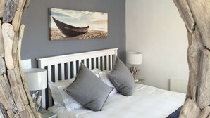 Ein Schlafzimmer mit Bett, Kissen, einem Nachttisch, einer Lampe und einem Gemälde von einem Boot an der Wand.