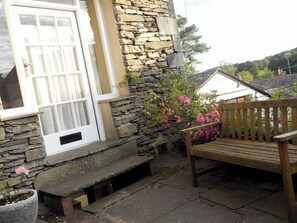 A stone building with a white door and windows, a wooden bench, and a potted plant with pink flowers.