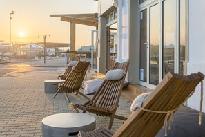 Buiten zitplaatsen met houten stoelen en kussens, een verhard terras en een gebouw met grote ramen.