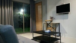 A modern living room with a sofa, a coffee table, a TV mounted on the wall, and a sliding glass door leading to an outdoor area. - Wara Boutique Hotel (Udon Thani)