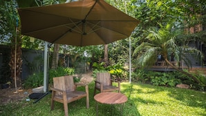 Un patio con dos sillas de madera y una mesa redonda pequeña, una sombrilla y abundante vegetación al fondo.
