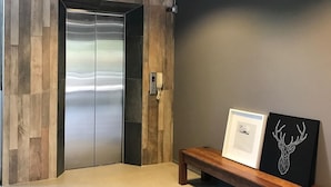 Un ascensor moderno con detalles metálicos, un panel de pared de madera, un banco con dos cuadros enmarcados y una lámpara colgante.
