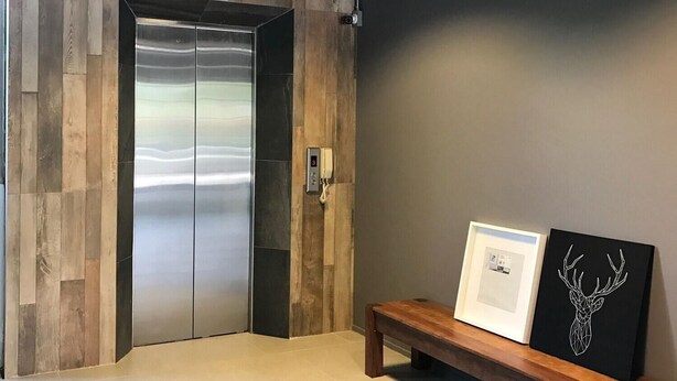 A modern elevator with a metallic finish, a wooden wall panel, a bench with two framed pictures, and a pendant light fixture. - Wara Boutique Hotel (Udon Thani)