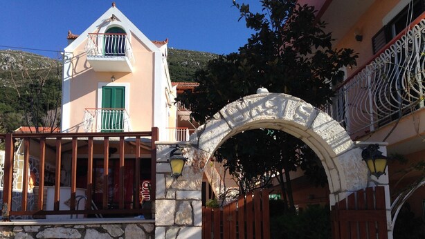 A stone gate with a metal archway leading to a building with a balcony and a green shuttered window. - Oasis Studios (Kefalonia)