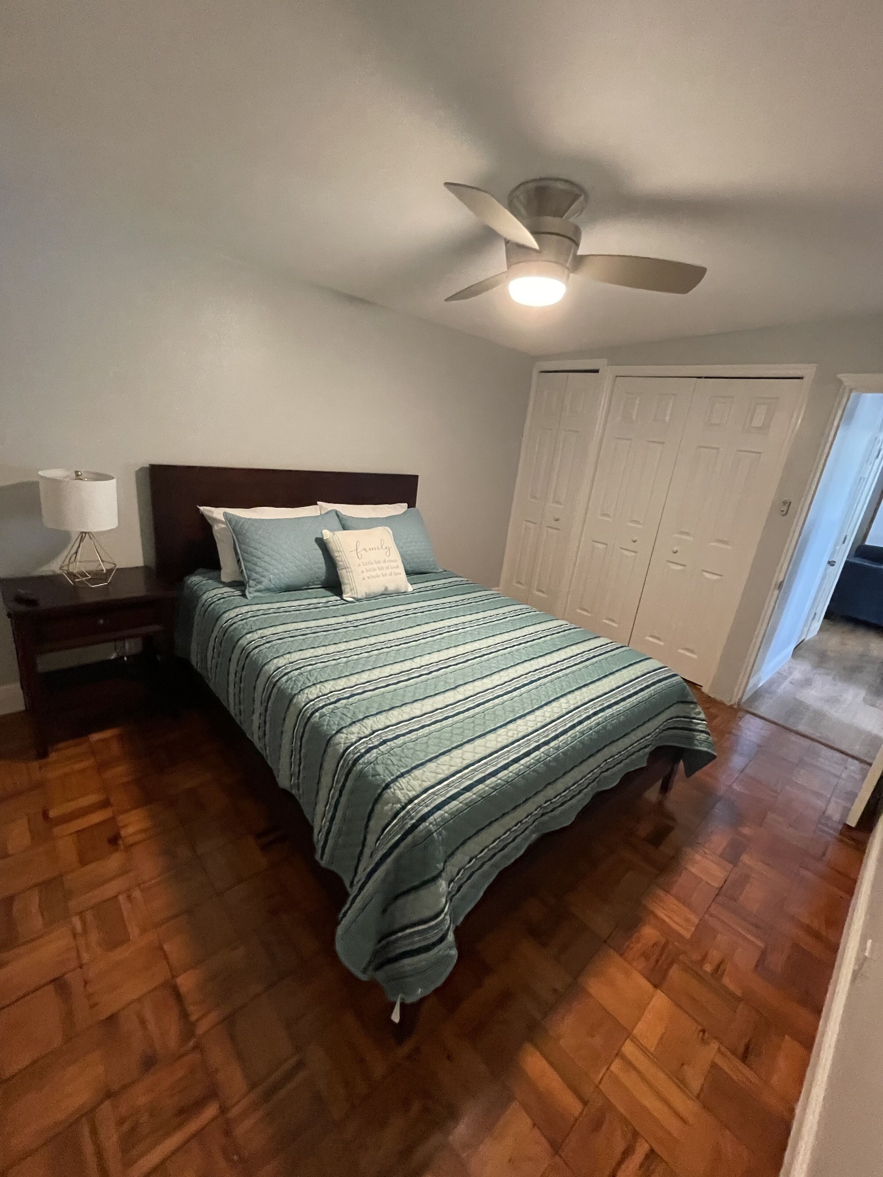 Un dormitorio con piso de madera, una cama con colcha a rayas, una mesita de noche con lámpara, un ventilador de techo y un conjunto de armarios blancos.