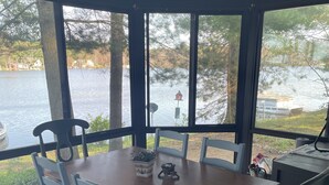 A dining area with a wooden table and chairs, large windows with a view of a lake and boats, and a small outdoor seating area with a table and chairs. - Northern Dream: All seasons lake house with nearby skiing, golfing and trails. (Gaylord)