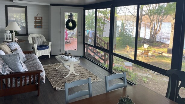 A cozy living room with a large window, a white lamp, a wicker chair, and a dining table. - Northern Dream: All seasons lake house with nearby skiing, golfing and trails. (Gaylord)