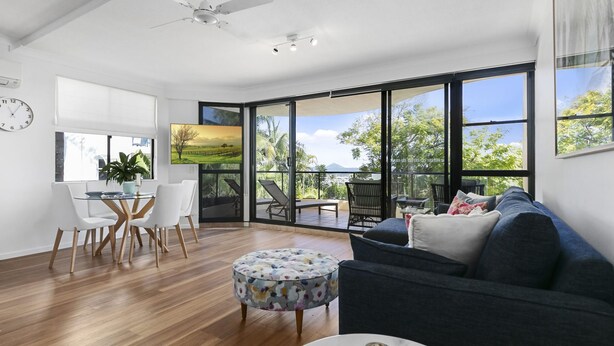 A spacious living area with a large window, a dining table with chairs, a sofa, and a decorative ottoman.