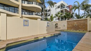 A swimming pool with a tiled deck, surrounded by a beige wall with decorative tiles and a small window. - Belle de Jour 1 (Noosa Heads)