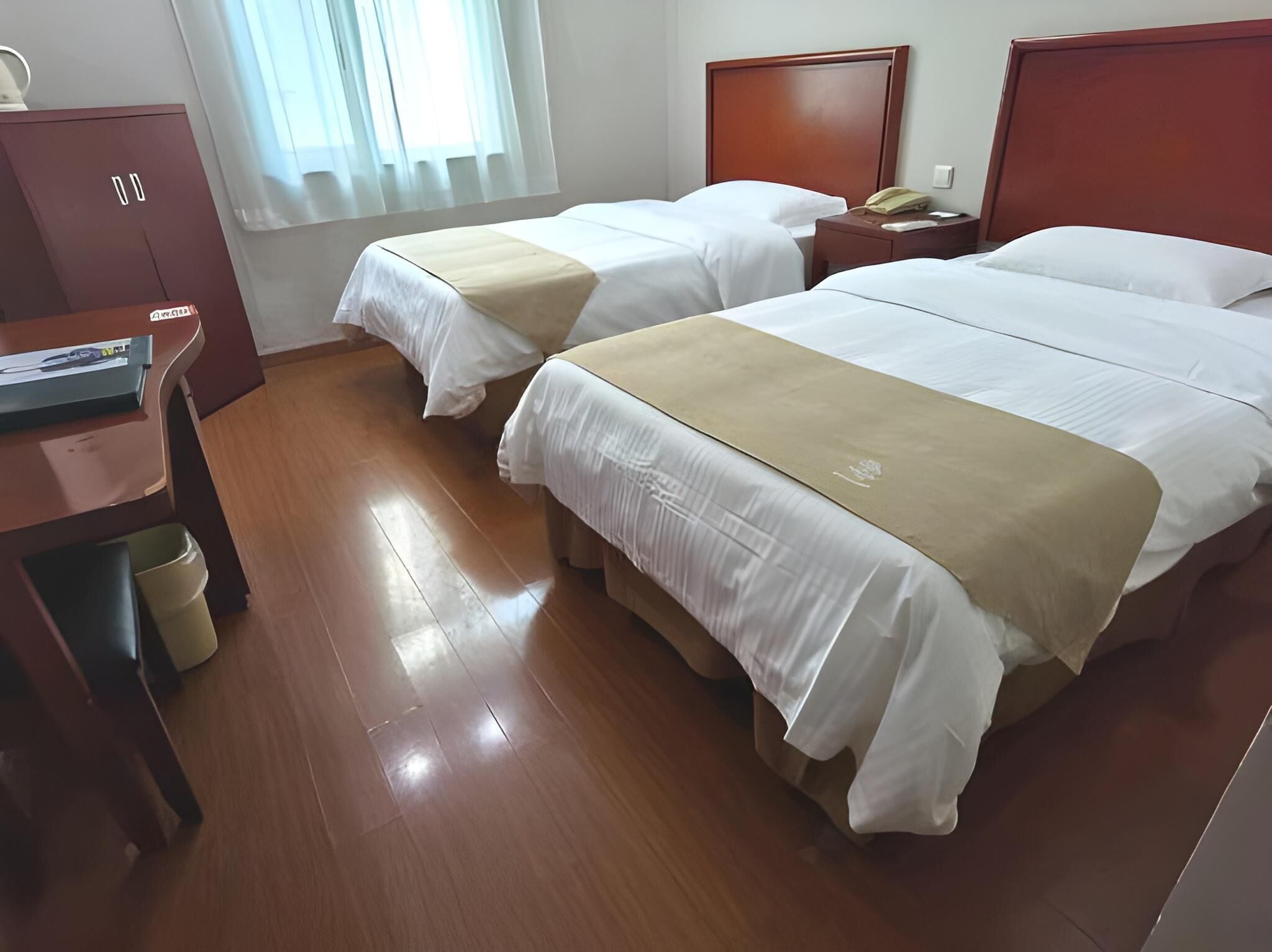 A hotel room with two beds, wooden flooring, a desk with a computer, a telephone, and a window with curtains.