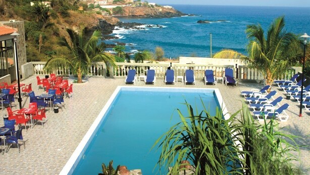 Area piscina con poltrone blu e rosse, vista libera sull'oceano e un pendio con edifici sullo sfondo.
