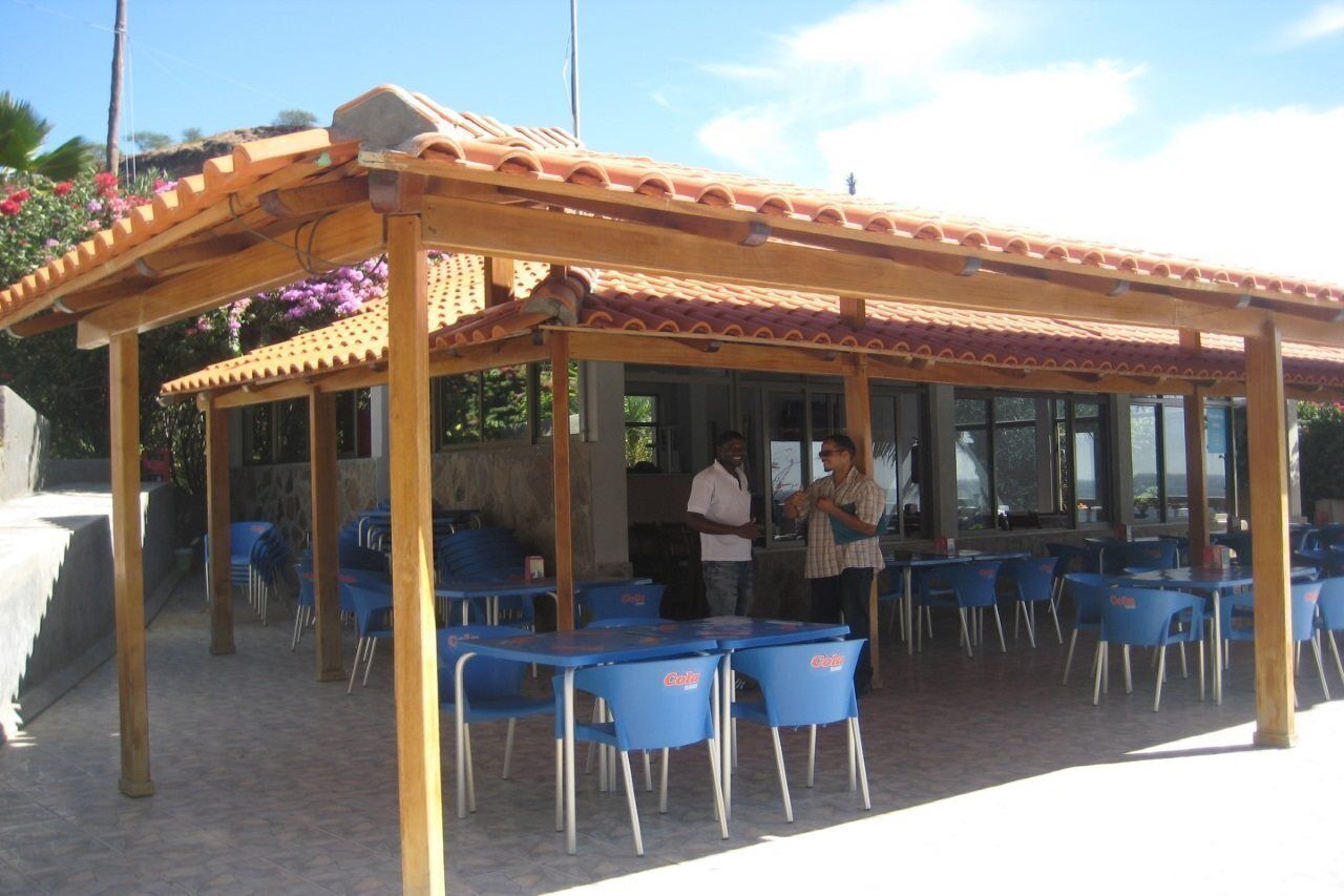 Pátio externo com pérgola de madeira, cadeiras e mesas de plástico azul.