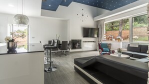 Una sala moderna con un amplio sofá seccional negro, un comedor con taburetes de bar, una cocina con horno empotrado y un gran ventanal con vistas al exterior.