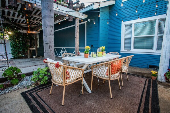 An outdoor patio with a white table and wicker chairs, surrounded by a blue building and string lights.