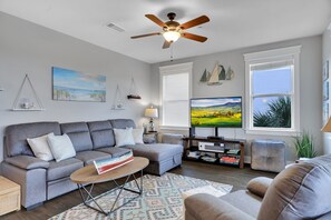 A living room with a grey sofa, a coffee table, a TV, and a ceiling fan. - Family sized Condo with bay views AND gulf views. Short walk to the beach! (Galveston)