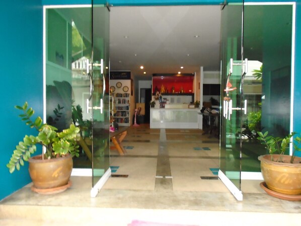 A modern reception area with glass doors, potted plants, a reception desk, and a visible kitchen area in the background.