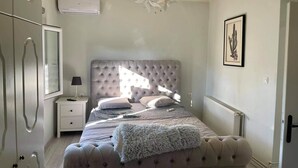 A bedroom with a tufted headboard, a large window, a radiator, a nightstand, a lamp, and a framed picture on the wall.