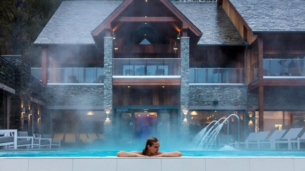 A person relaxing in a hot tub in front of a large, modern building with a stone facade and multiple balconies. - Lodore Falls Hotel (Keswick)