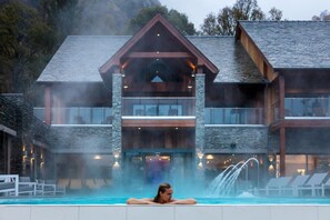 A person relaxing in a hot tub in front of a large, modern building with a stone facade and multiple balconies. - Lodore Falls Hotel (Keswick)